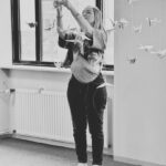 A woman stands indoors, reaching up to adjust or hang paper cranes suspended from strings. She is surrounded by the hanging origami birds and is facing slightly to the right. The photo is in black and white.