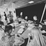 A group of people sit around a table covered with papers and drinks, engaged in discussion. Two others stand nearby, smiling and observing. The setting appears to be an office or conference room. The image is in black and white.