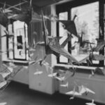 Black and white photo of numerous paper cranes made from written-on paper, hanging from the ceiling in front of large windows, creating a sense of movement and lightness in the room.