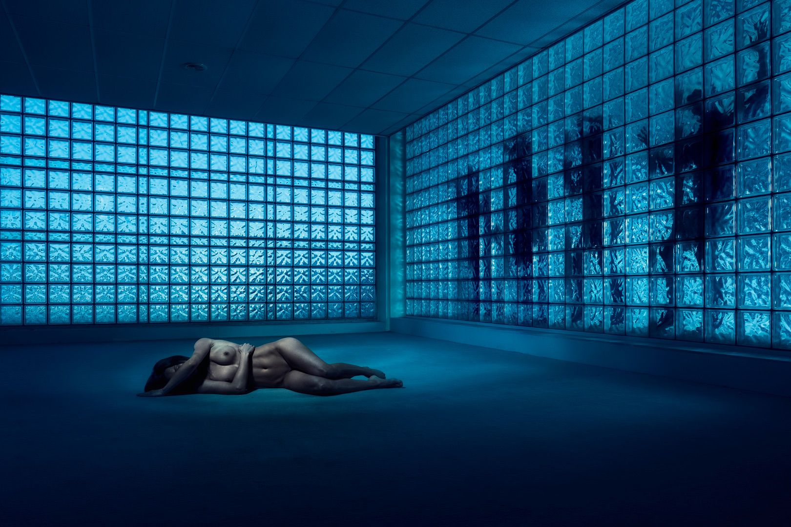 A nude person lies curled on the floor of an empty, blue-lit room with glass block walls; shadowy figures loom behind the glass, creating an eerie atmosphere perfect for a hauntingly memorable portfolio piece.