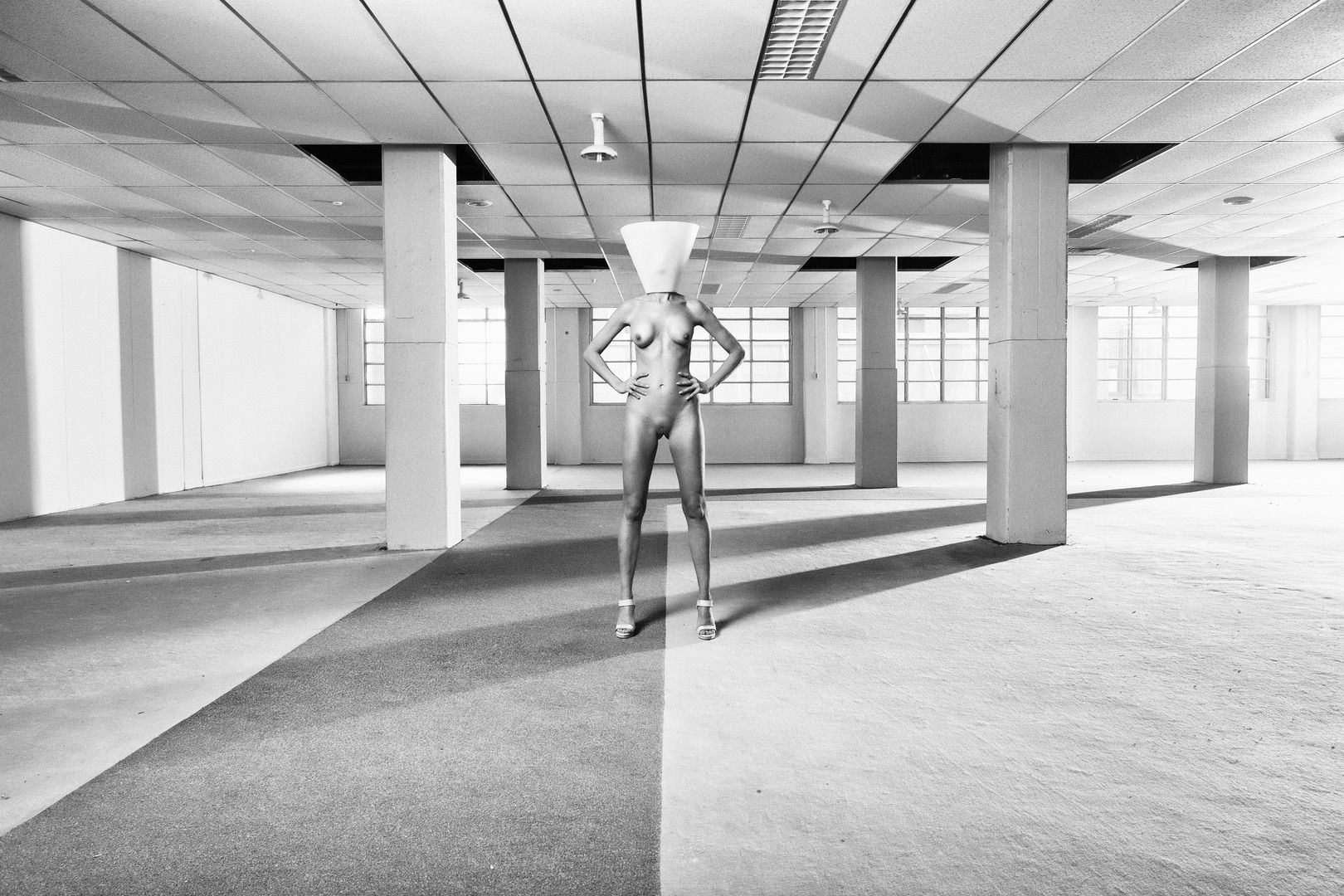 A striking black and white portfolio image features a person in high heels and a cone-shaped headpiece, standing confidently with hands on hips in an empty industrial-style room, pillars and tiled ceiling illuminated by window light.