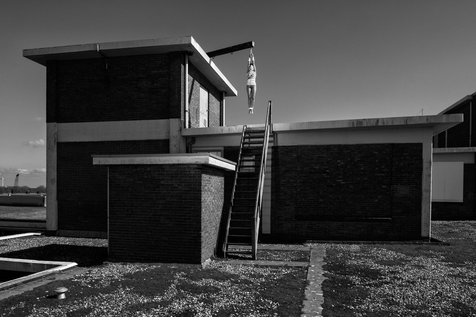A black and white photo for your portfolio: a brick building with an outdoor staircase. At the top, a person hangs by their arms from a metal beam, legs dangling above the stairs under a clear sky.