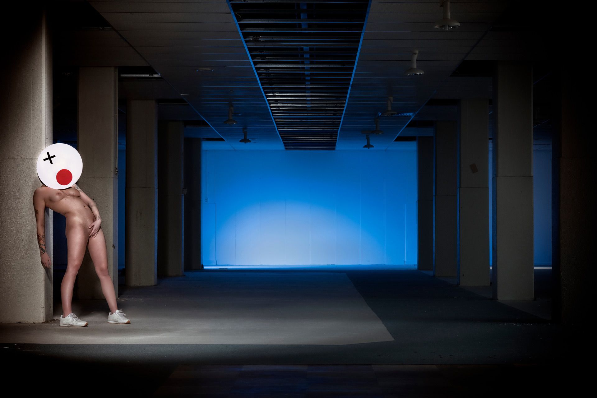In a dimly lit, empty building with blue light, someone nude except for white sneakers and a playful cartoon mask poses by a column—forgetting eyeliner but making up for it—presenting "Time flies when I masturbate.