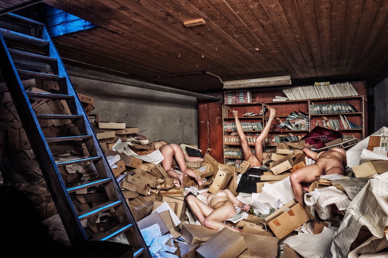 A cluttered room filled with scattered boxes, papers, and books—an unexpected portfolio of chaos—features several nude people partially buried among the mess, while a blue-tinged wooden ladder leans against the wall on the left.