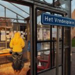 A person with long blonde hair stands in a glass structure, reading display boards. A blue sign on the door reads "Het Vredesplein." The space is warmly lit and decorated with a carpet and furniture.